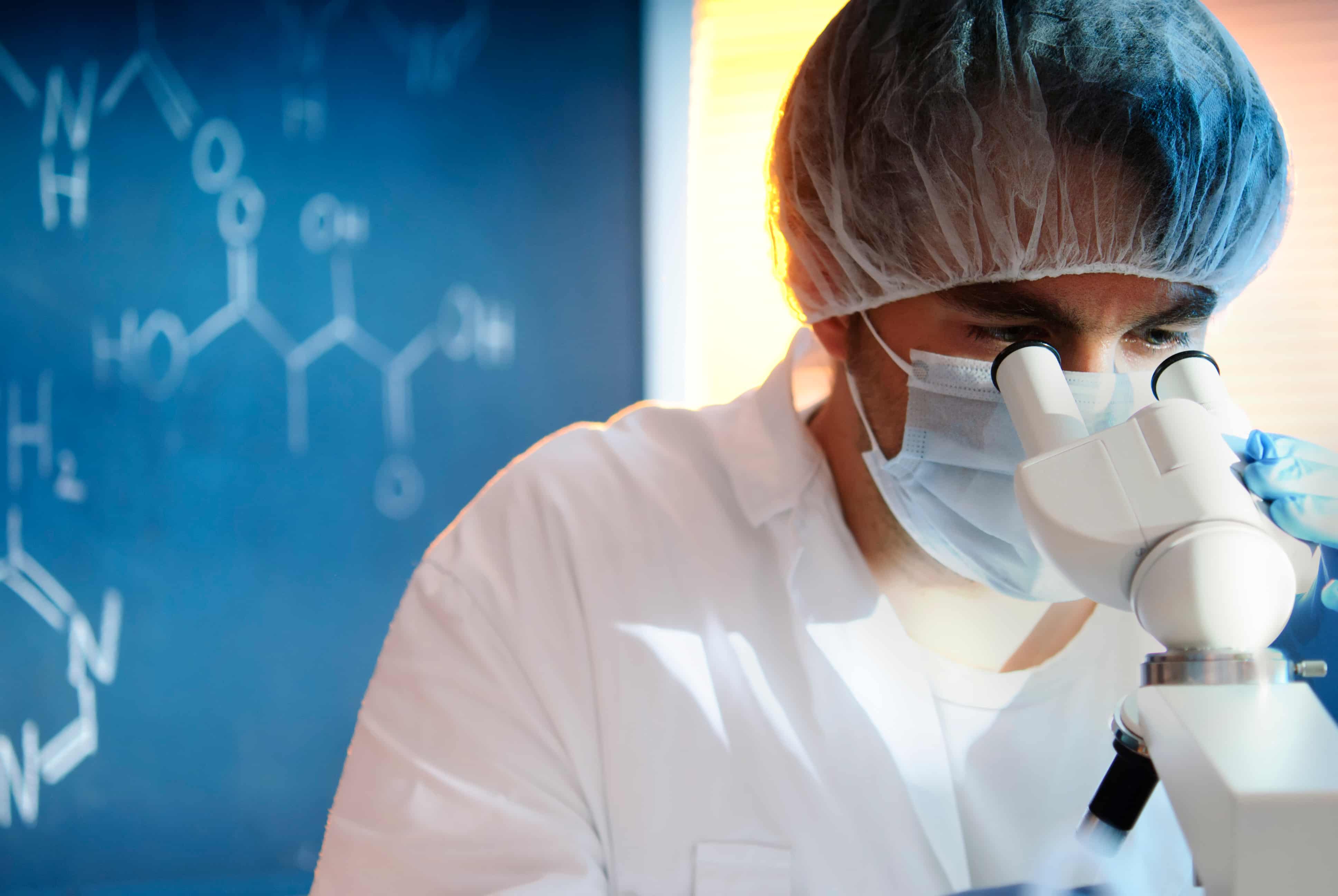 Laborwissenschaftler mit Haarnetz und Schutzmaske blickt konzentriert in ein Mikroskop