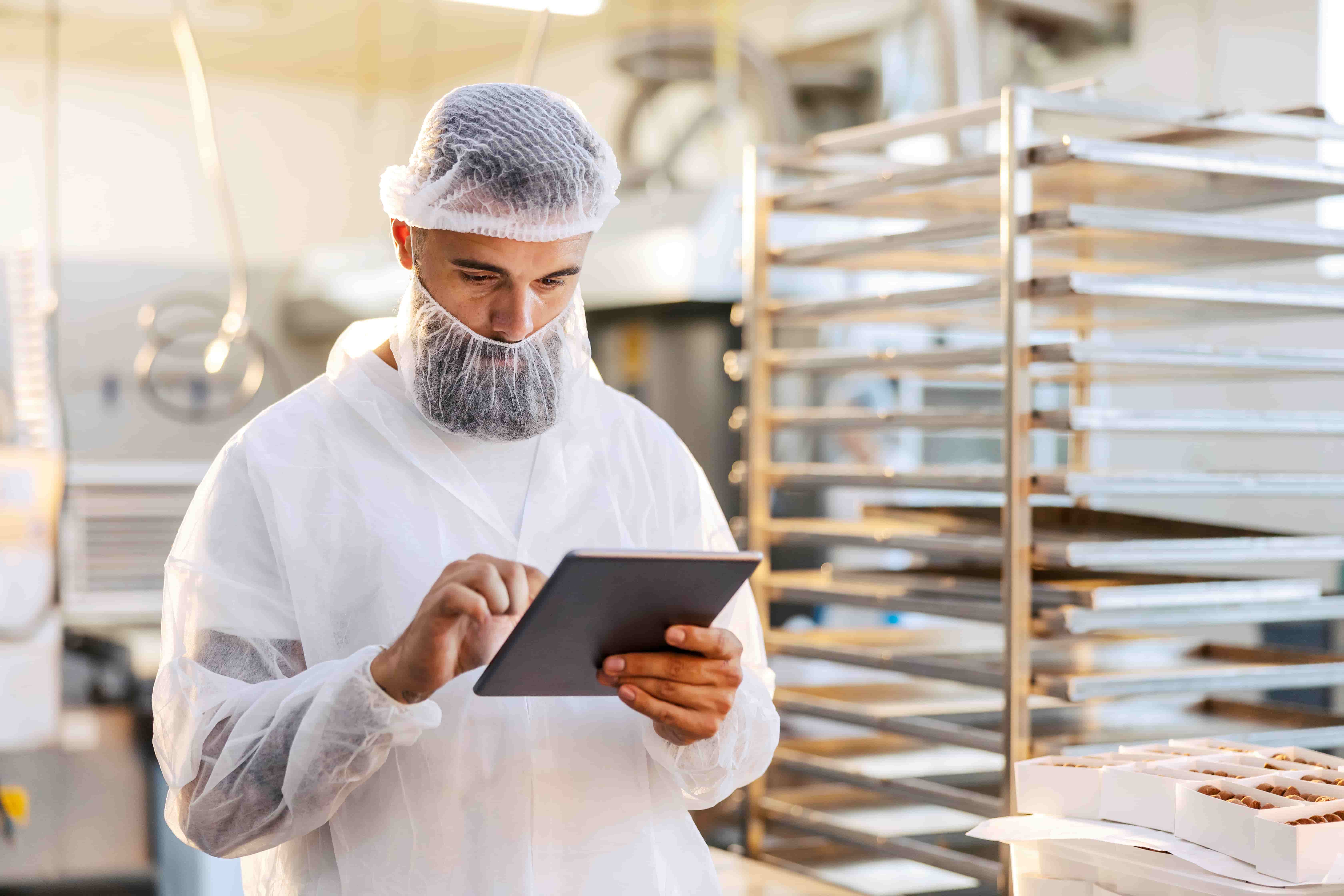 Mitarbeiter mit Bartschutz und Haarnetz in der Lebensmittelproduktion nutzt Tablet